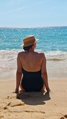 A back view of a stylish woman in black one-piece swimsuit and straw hat, sitting on golden sandy beach, looking at the turquoise ocean under clear blue sky. Ideal for wellness and travel imagery.