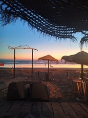 Tranquil beach sunset with straw umbrellas, lounge chairs on sandy shore, calm ocean waves, silhouettes of people and dog walking. Warm pink-orange sky.Perfect for travel, leisure, and paradise themes