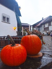 Cozy autumn scene in a Swiss village. Vibrant orange pumpkins on a wet stone ledge. Old white houses, warm lantern light, cobblestone path. Cozy village pictures. Halloween scene, decor. Fall getaway
