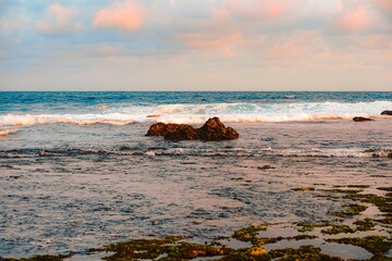 Vast, calm ocean view at low tide with white waves cresting over a central rock, reflecting the pink and blue hues of the evening sky.