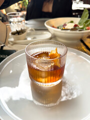 Glass of amber drink with ice and orange peel served on a reflective plate. Elegant cocktail presentation in a fine dining atmosphere.