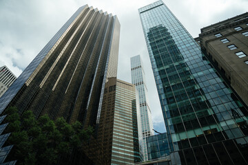 A towering view of glass and steel skyscrapers in Midtown Manhattan, New York City. The architecture reflects light through the cloudy sky, showcasing the vertical power of the urban skyline.