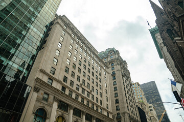 A mix of stone facades and glass buildings rises into a cloudy New York sky. Reflections and textures highlight the contrast between old and new architecture.