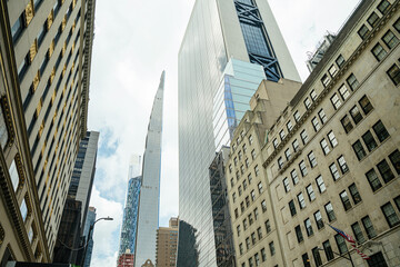 A mix of modern skyscrapers and historic architecture lines a New York street. The American flag waves in the foreground while sharp towers rise high into the sky.