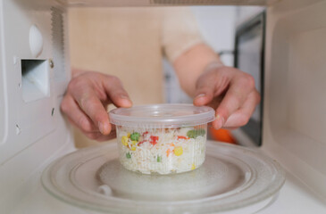 A close-up view from inside a microwave as a person places a plastic container of frozen rice and vegetables onto the glass turntable.