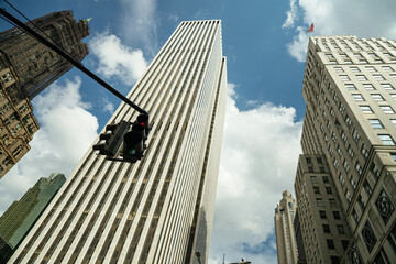 Upward view of Manhattan skyscrapers under a bright blue sky with scattered clouds. A red traffic light is suspended in the foreground, adding contrast to the towering buildings.