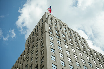 Low-angle view of a beige office building with many square windows and an American flag waving on top. The bright sky with scattered clouds highlights the sharp architectural lines of the structure.