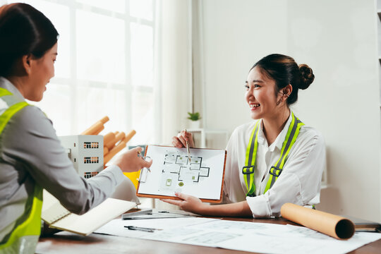 Two happy female architects wearing safety vests are discussing a project comparing blueprints and a scale model in their office