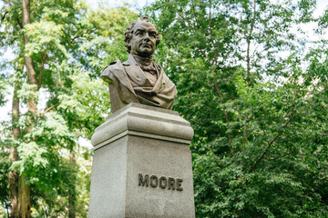 A detailed close-up view of the bronze bust of Thomas Moore in Central Park. The statue rests atop a granite pedestal surrounded by lush green trees and soft sunlight.