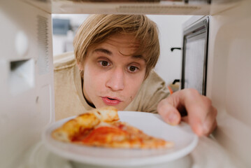 Young man looking into a microwave while placing a plate with a slice of pizza inside. Meal preparation, bachelor life, convenience food.