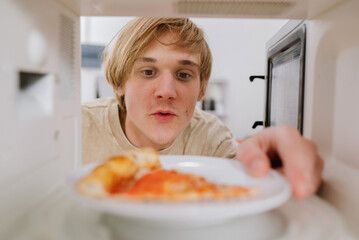 Young man looking into a microwave while placing a plate with a slice of pizza inside. Meal preparation, bachelor life, convenience food.