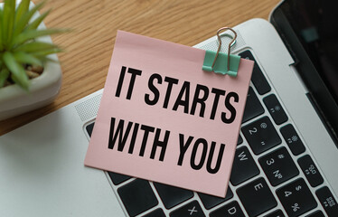 A pink piece of paper with the words It Starts With You written on it is sitting on top of a laptop keyboard
