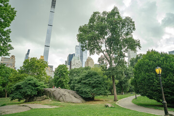 A winding path and rocky hill are surrounded by green trees in Central Park, New York City. Tall modern skyscrapers and a vintage lamp rise in the background under a cloudy sky.