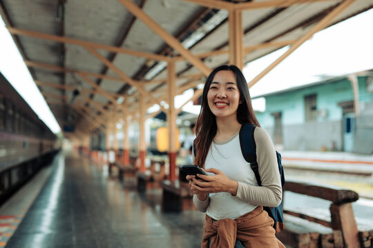 Asian female traveler is smiling and using smartphone while waiting for train at railway station