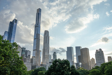 Tall skyscrapers dominate the skyline of Midtown Manhattan, rising above the trees of Central Park. The glass buildings reflect the daylight under a dramatic blue sky with scattered clouds.