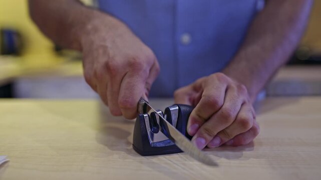 Close-up of hands sharpening a kitchen knife using a manual sharpener on a wooden countertop. The video captures the clear, crisp sounds of the blade being honed, highlighting a domestic kitchen task