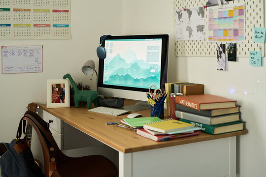 Organized student desk featuring desktop computer, large stack of textbooks, stationery, framed family photo, dinosaur figurine, wall calendars, motivational notes, world maps