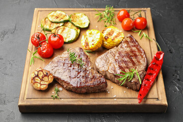 Delicious grilled beef steaks with herbs and vegetables on dark textured table, closeup