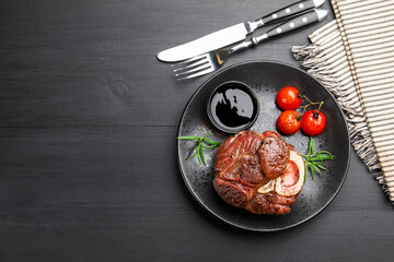 Tasty grilled beef steak served with rosemary, sauce, tomatoes and cutlery on black wooden table, flat lay. Space for text