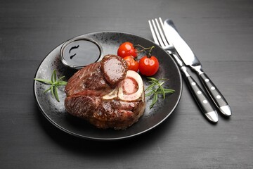 Tasty grilled beef steak served with rosemary, sauce, tomatoes and cutlery on black wooden table