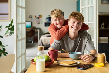 Caucasian teenage brothers sitting at kitchen table, older brother eating breakfast while younger brother hugging him from behind, both smiling, smartphone and food on table