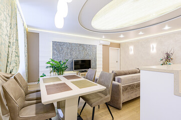 A modern living space with a dining table, upholstered chairs, a sleek TV setup, and stylish lighting. The room features soft colors and contemporary design