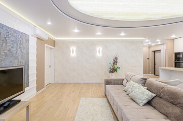 A bright living room features a soft beige sofa, wooden floors, and a decorative ceiling with recessed lighting, creating a modern and inviting space