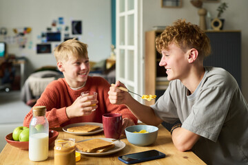 Two Caucasian teenage boys sitting at table eating breakfast together, one holding glass of juice while other eating cereal with spoon, both smiling and interacting in casual home setting © pressmaster