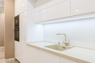 kitchen features sleek white cabinets, light-toned countertop with matching sink and faucet. backdrop is textured white tile wall with under-cabinet lighting. Built in black ovens provide contrast