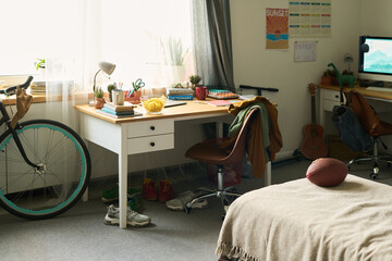 Modern bedroom workspace showing organized desk with books, potted plants, bicycle, sneakers, football on bed, computer monitor, and guitar, sunlight streaming through window