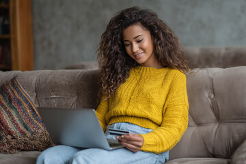 Online Shopping Delight: A young woman, nestled on a cozy sofa, is absorbed in the seamless world of online shopping, holding her credit card and browsing on her laptop.