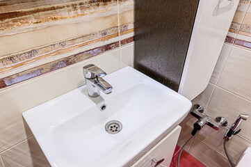 A small, modern bathroom with patterned beige tiles, a white toilet and sink, and a red-tiled shower stall visible through a glass door. Bright lighting emphasizes cleanliness