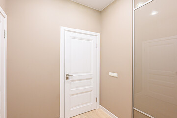 A bright hallway interior featuring a white paneled door, light beige walls, and a mirrored wardrobe. A clean and simple design conveys a sense of order