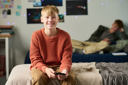 Portrait of Caucasian teenage boy sitting on bed holding smartphone smiling at camera, with another Caucasian teenage boy reclining on bed in background using digital tablet