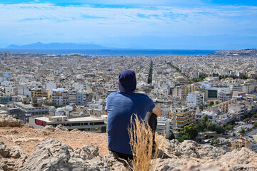 Photo of an unrecognizable person sitting at on a tock at the Filopappou hill in Athens and watching the southern suburbs of the city. Blue sky with clouds and the sea at the horizon.