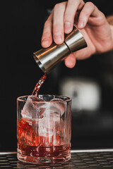 Bartender pours red cocktail from jigger into glass with ice cubes. Close-up shot, dark background. Professional mixology, drink preparation, nightlife concept.