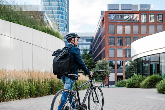 City commuter walking with bike to office, wearing backpack and helmet.