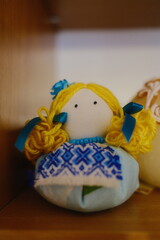 Doll wearing a blue and white patterned dress sits upright on a brown wooden shelf.