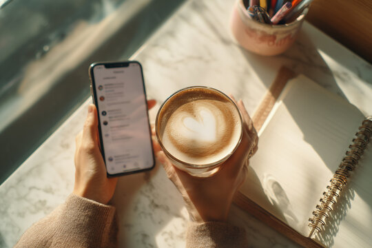 Cozy morning ritual: hands holding a heart-shaped latte while scrolling a smartphone beside an open notebook on a sunlit marble workspace