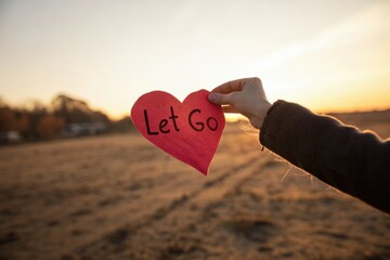Hand holding red heart with "Let Go" message outdoors at sunset