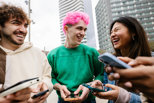 Diverse young friends having fun using mobile phones at city street. Millennial generation people laughing while watching social media content on cellphone app