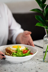 Top view of a breakfast plate with sunny-side-up egg on toast, fresh salad with cherry tomatoes, hand reaching for food, minimal table setting with vase and green plant.