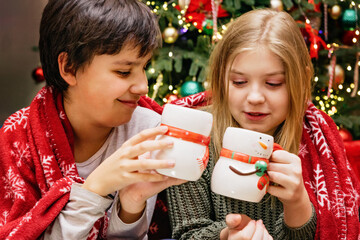 Christmas and New Year holiday celebration concept. Two children wrapped in red blankets holding festive snowman mugs with hot drinks in front of decorated Christmas tree and holiday lights