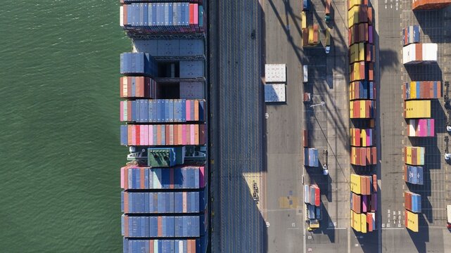 Oakland, United States - 02 September 2025: Aerial view of stacked shipping containers bursting with vibrant colors contrast against the tranquil, green water, forming a geometric tapestry.