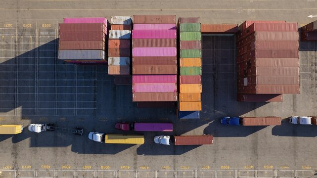 Oakland, United States - 02 September 2025: Aerial view of stacked shipping containers casting long shadows, next to trucks ready for transport, a symphony of industrial colors.