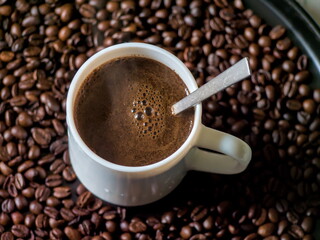 delicious-looking cup of freshly brewed black coffee sits in a bed of roasted coffee beans. A small spoon is in the cup, ready for stirring, creating a perfect scene for a morning coffee break
