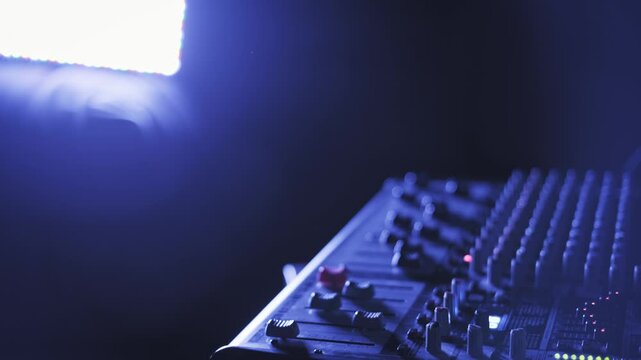 Panning shot across audio mixer console in a dark space, showing faders and knobs illuminated by cool blue light, with a bright LED panel and lens flare visible in the background for atmosphere.