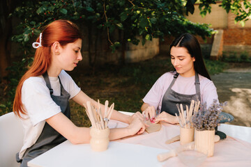 Two women enjoy a pottery class outdoors, shaping clay with smiles and creativity. 2 Girls sculpt clay in an outdoor workshop. Clay art workshop outdoors.