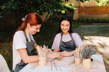 2 Girls sculpt clay in an outdoor workshop. Clay art workshop outdoors. Two women enjoy a pottery class outdoors, shaping clay with smiles and creativity.