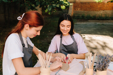 Two women enjoy a pottery class outdoors, shaping clay with smiles and creativity. 2 Girls sculpt clay in an outdoor workshop. Clay art workshop outdoors.